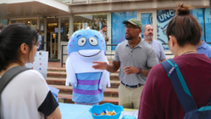 Mr. Phish costumed creature observes Information Security Office staff member interact with students at the 2018 Tech Fair.