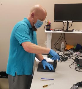 Wearing a mask and gloves, an employee uses a power screwdriver on a laptop