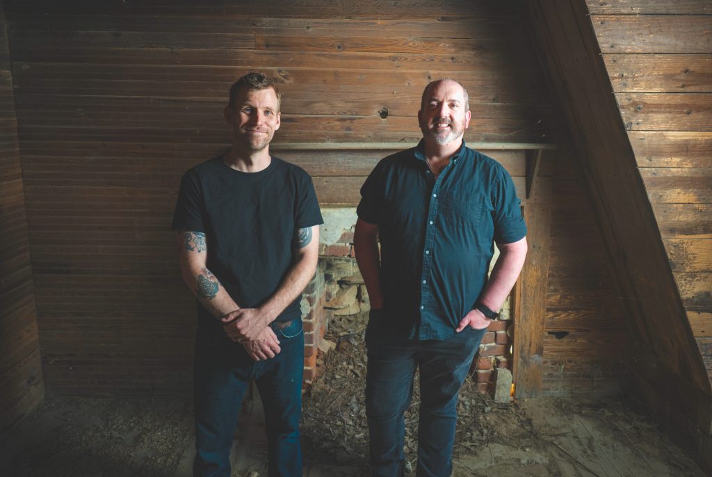 Charlie Mewshaw and Michael Williams pose in front of a crumbling fireplace inside an old, abandoned wood-paneled room.