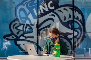 A student uses a laptop on a bistro table in front of student stores, where a decal of the Ramses mascot fills the windows
