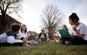 Five students sit in a semicircle on the grass in Polk Place. Each has a laptop on the ground or on their lap