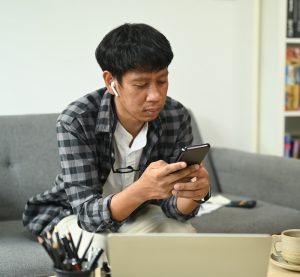 A man sits on a couch. He's wearing airpods and looking at his smartphone. His laptop is on open on the coffee table in front of him.