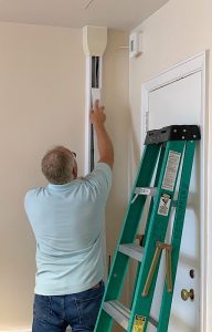 A man stands on a ladder as he installs an access point on the wall near the ceiling.
