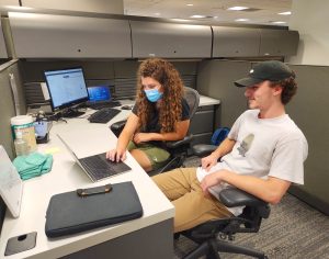A student worker types on a laptop while her client watches as he sits next to her at a desk.
