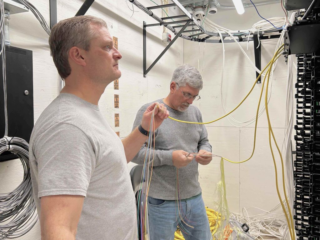 Danny Maddox and Kevin Wicker hold and connect fiber cables in a telecom closet.