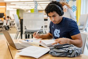 A student, wearing headphones, compares his notes to his textbook while also using his laptop