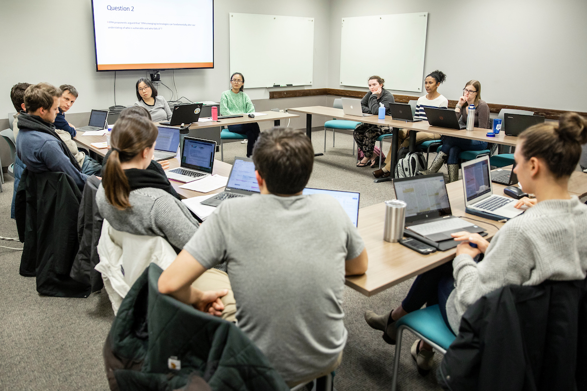 Students sit at a tables configured into a square while they listen to their professor. Each student has an open laptop in front of them.