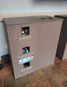 A tall cabinet has three vertical compartments, one labeled e-waste, another for plastic film and a third for batteries