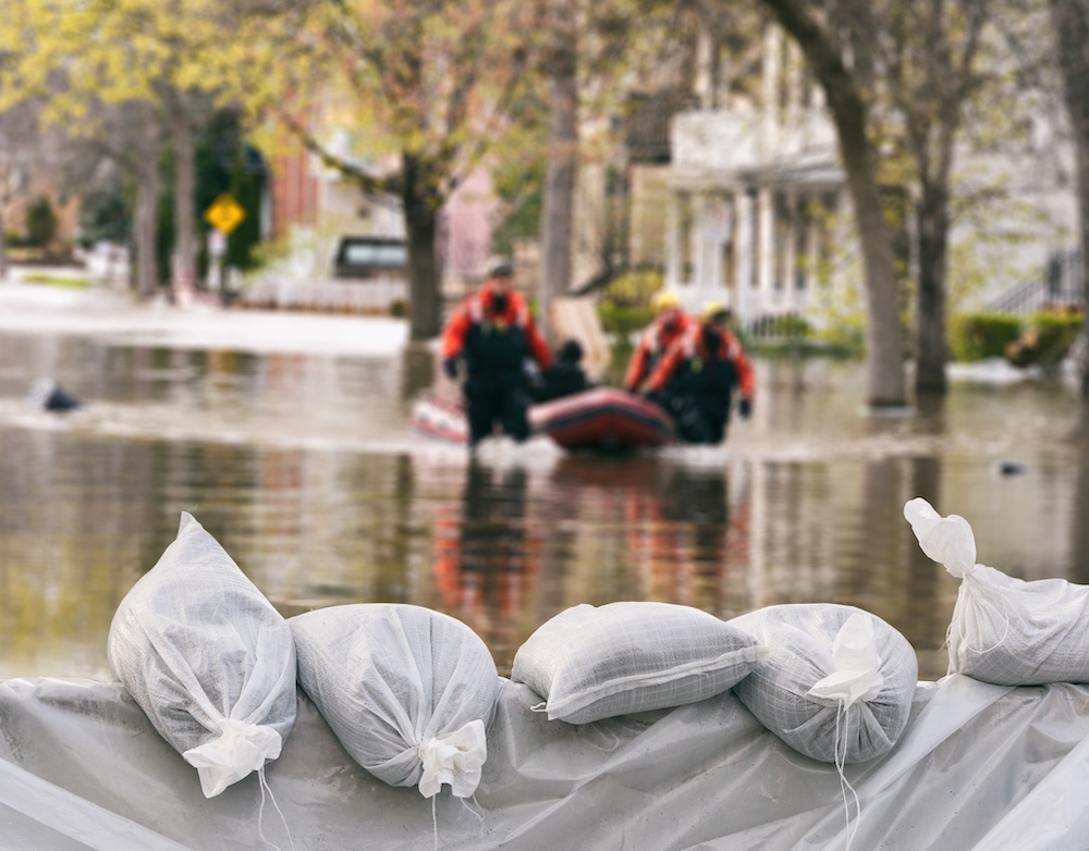 In the foreground are flood protection sandbags. In the background, a water rescue happens on a flooded street