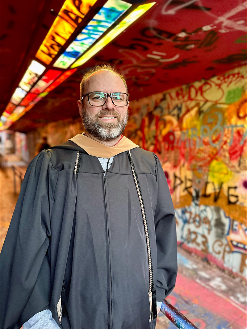 Phil Young, wearing graduation regalia, stands in the graffitied Free Expression Tunnel at NC State University