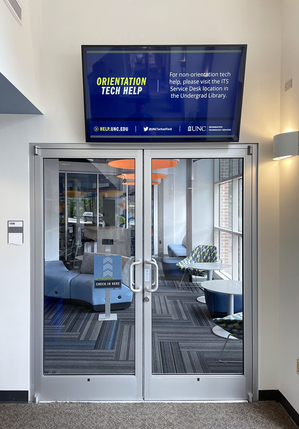 A glimpse through the glass doors from the Union into the Service Desk space. A screen over the doors reads Orientation Tech Help