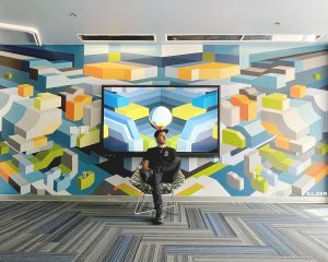 Artist ILL.DES sits in a chair in front of the completed mural