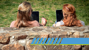Two young women sit with their backs to a stone wall on a sunny day on the quad. They're each holding a laptop