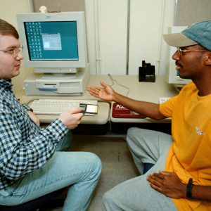 Two students sit in front of beige desktop computers. A Service Desk employee hands over a floppy disk