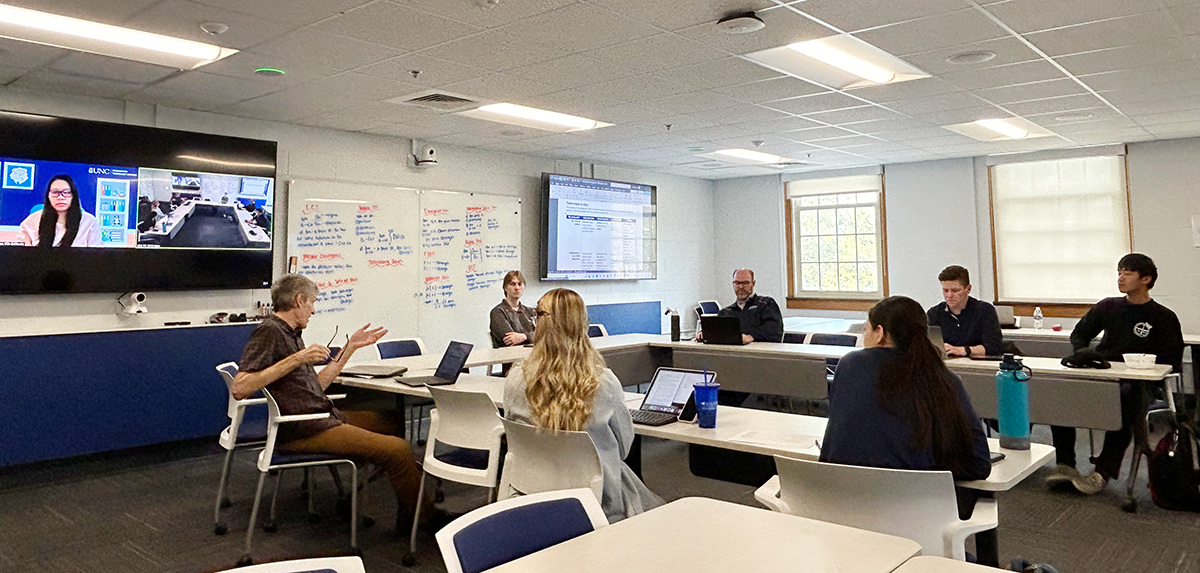 At a recent STC meeting, a group of students and staff advisors sit in a classroom in at tables in a semicircle. Bob Henshaw gestures and references a presentation.