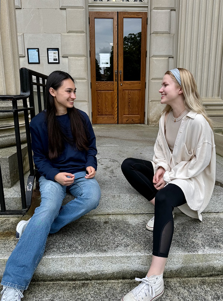 Two students smile at each other while sitting on building steps with a door and columns behind them.