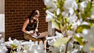 On UNC's campus, a woman sits against a brick wall and uses her laptop. She's framed by blooming dogwood flowers